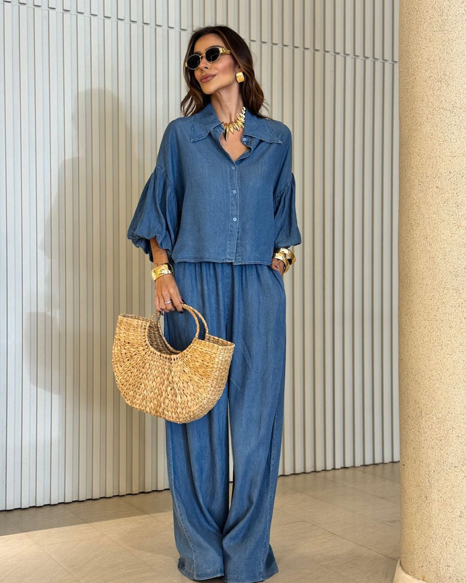 Modelo usando conjunto all jeans azul: blusa com manga balonê e botões frontais, calça pantalona longa com elástico na cintura.